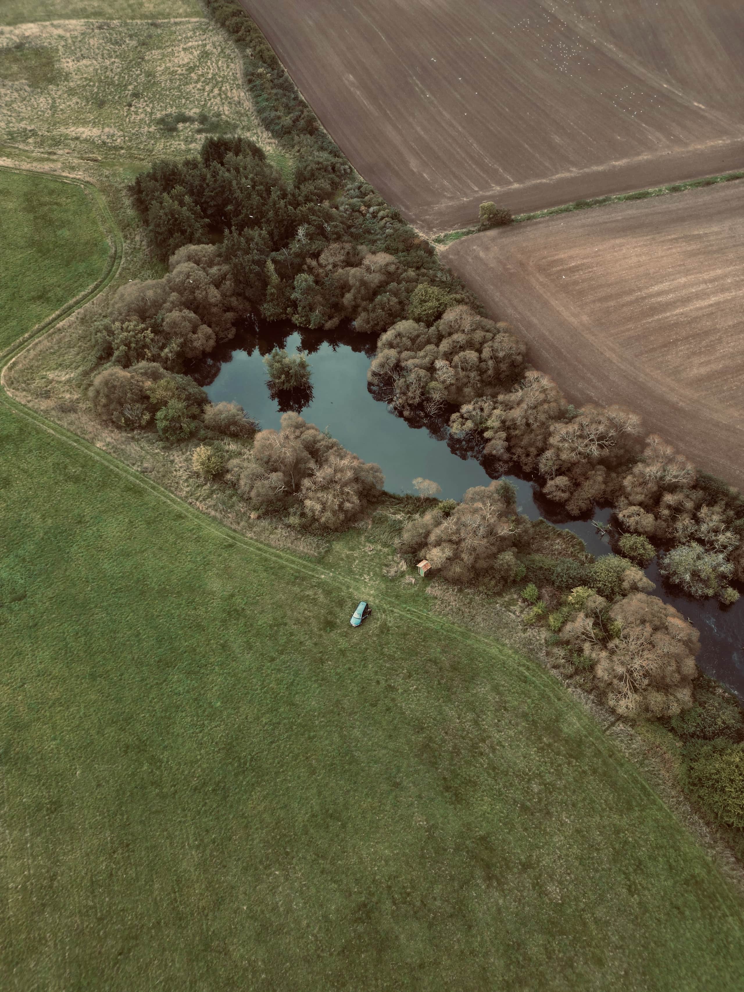 Luftfoto af en lille sø omgivet af træer og marker med en bil på græsset.